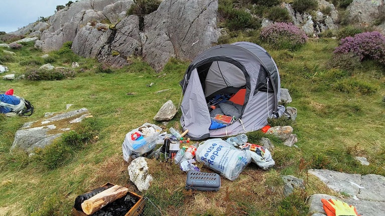 A grey tent situated on moorland in front of a rocky outcrop is surrounded by bags of rubbish and a used fire pit.